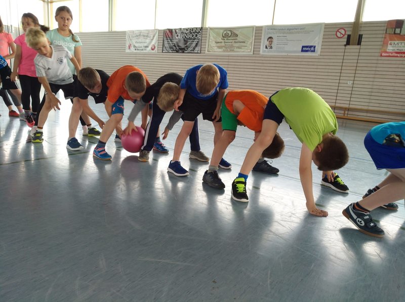 Grundschulkinder stehen in einer Turnhalle gebückt in einer Reihe und geben einen Ball durch ihre Beine weiter